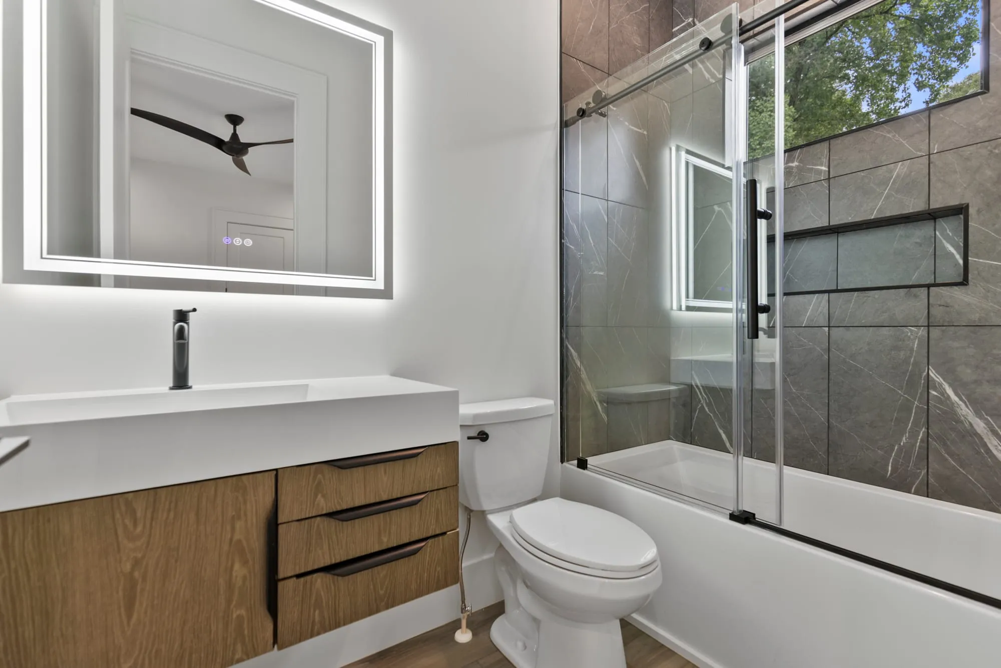 Remodeled bathroom with a backlit mirror, floating wood vanity and marble-tile shower